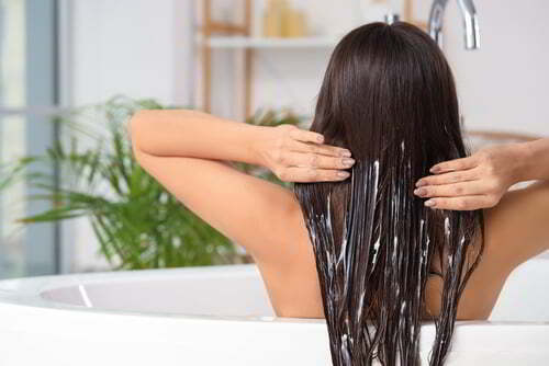 צילום אחורי על אישה צעירה מורחת מרכך או מסכה לשיער בחדר אמבטיה